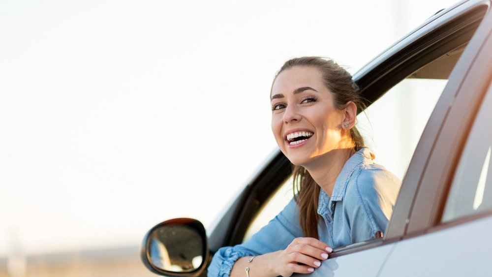A Woman smiles out of the drivers door windows of her car.